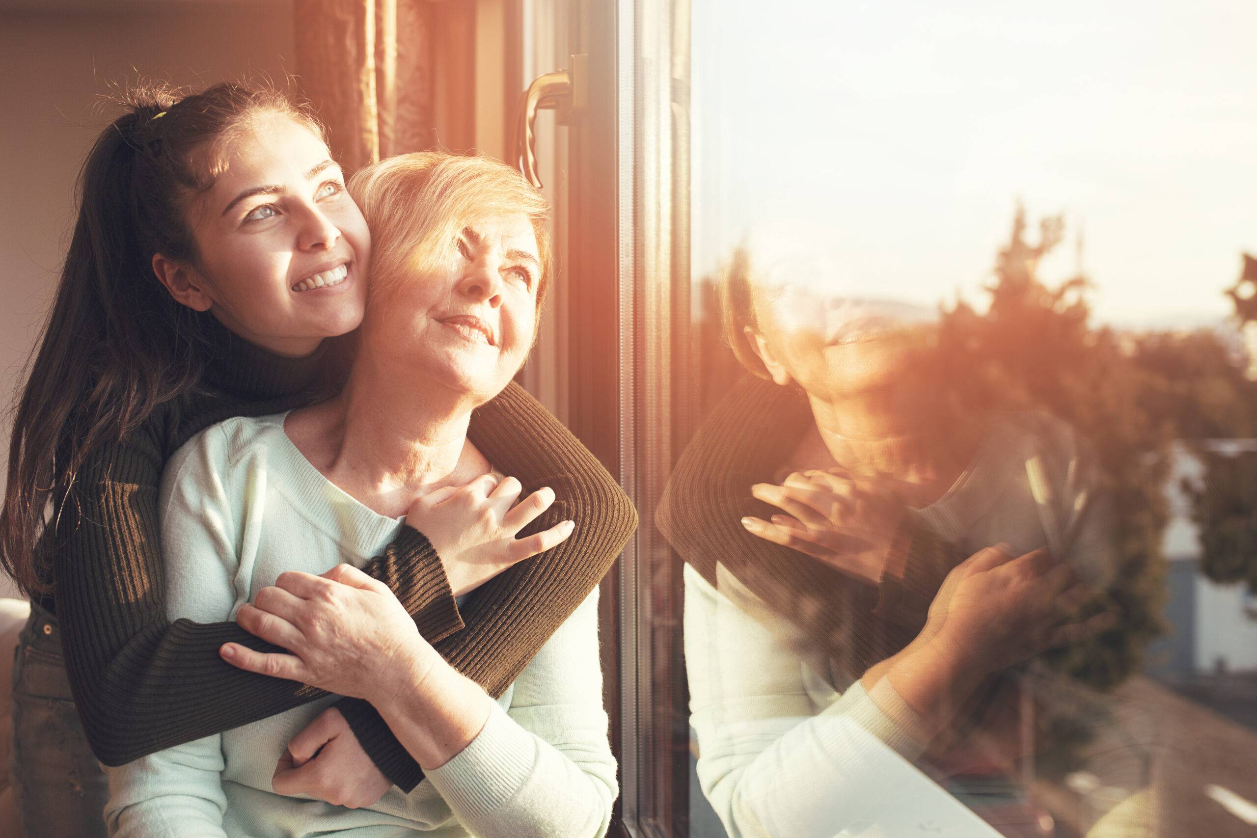 Young person hugging an older woman, looking out the window with the sun shining on their smiling faces.